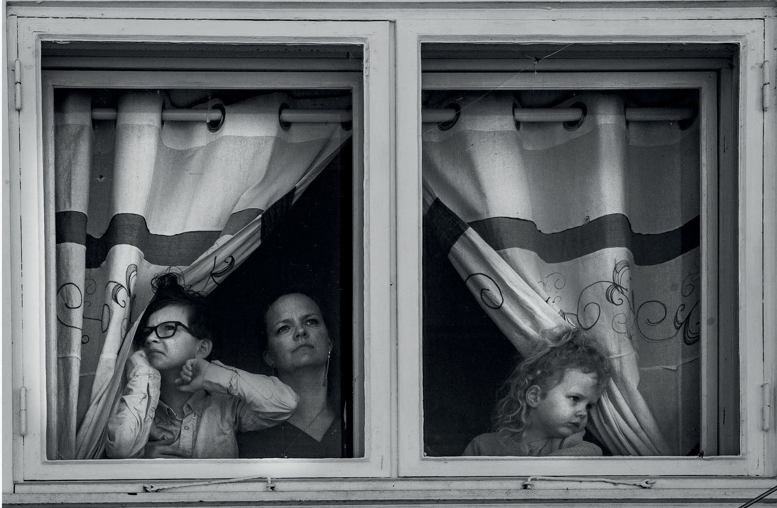 Two women and a child at a flat window, behind half-drawn curtains.