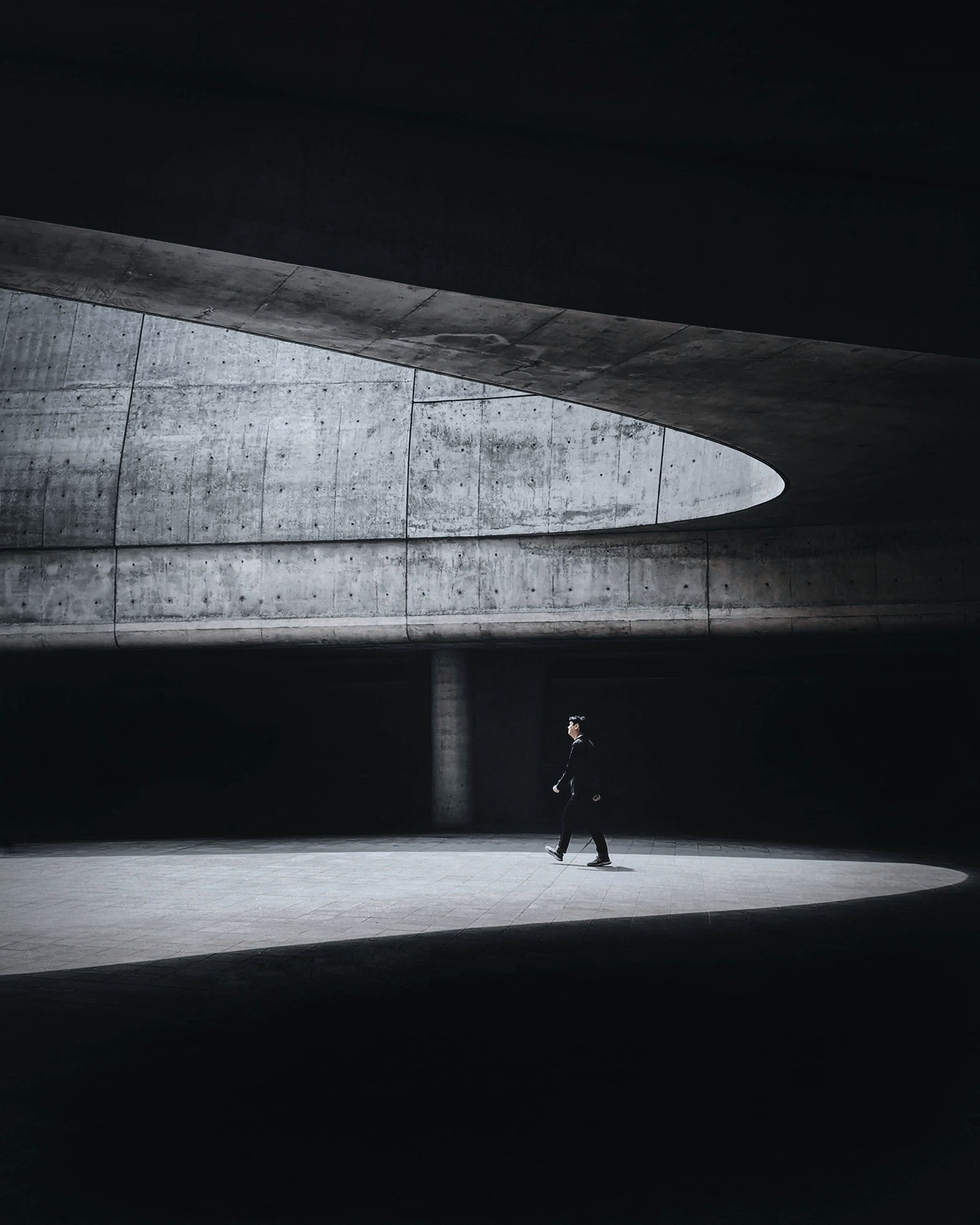 A single figure crossing a pool of light in a dark concrete hall, a curved opening above.
