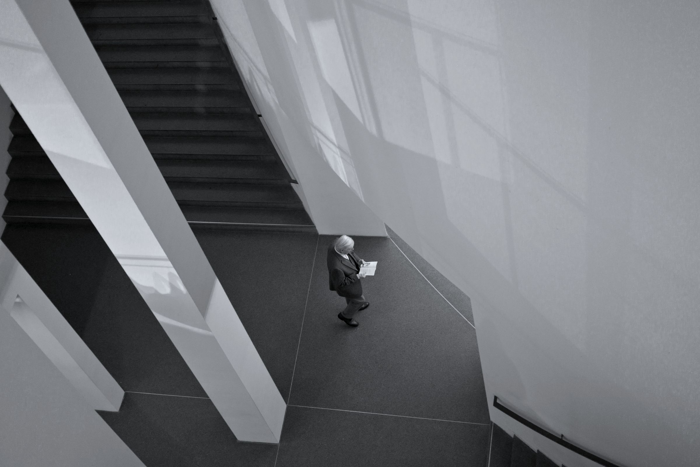 Seen from above, a person reading a sheet of paper at the foot of a pale staircase.