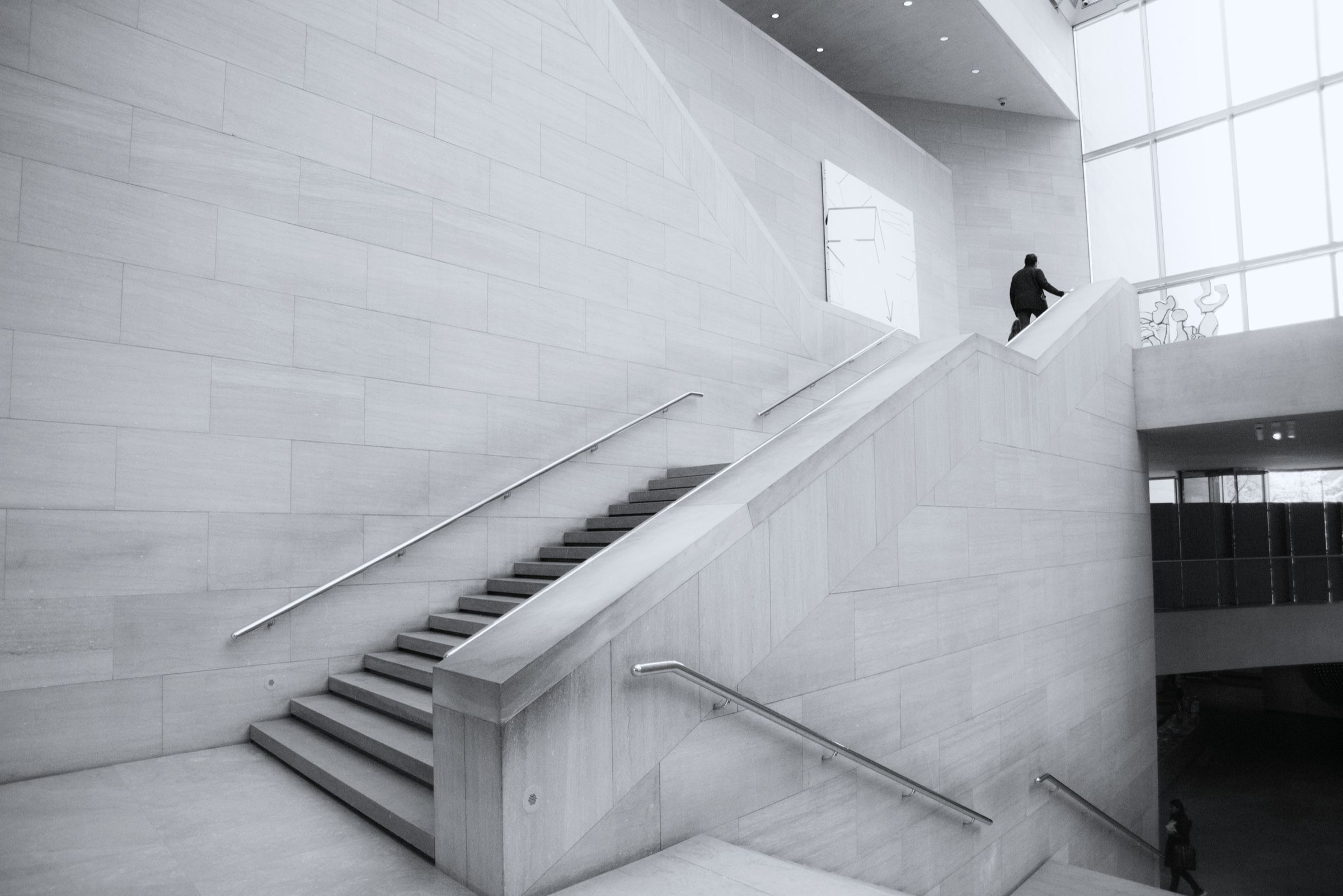 White stairwell in daylight, with a single person on the upper flight.