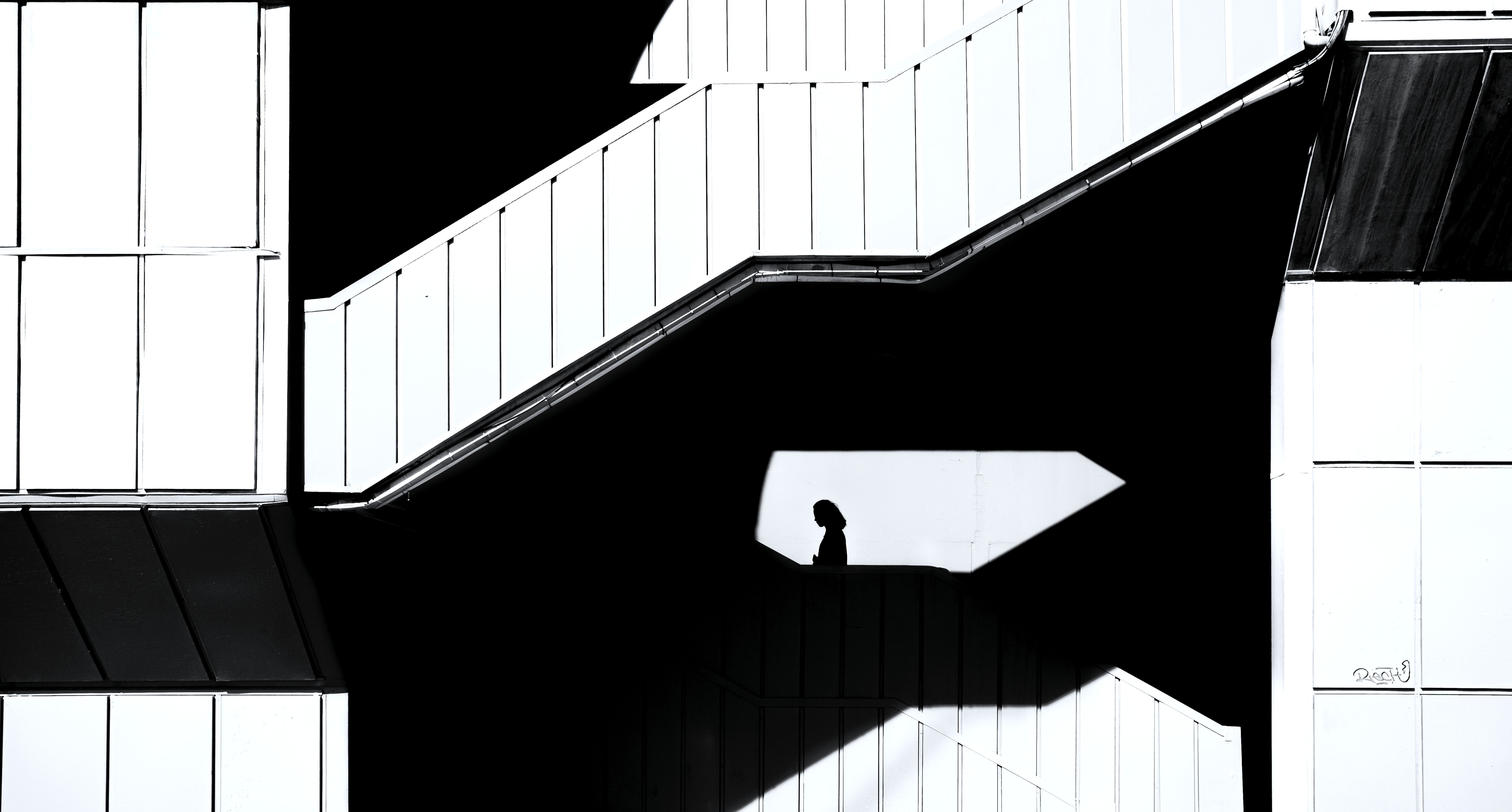 A figure in silhouette on an external stair, in a patch of sunlight on a white facade.