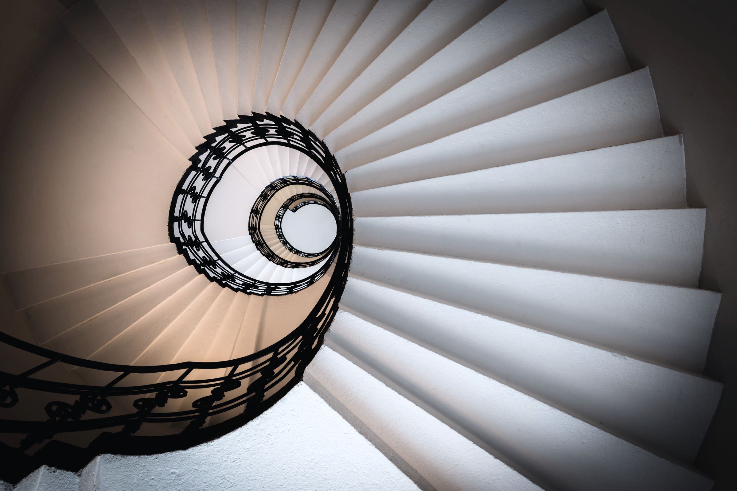 A spiral stair seen from below, its treads winding up to a small oculus of light.