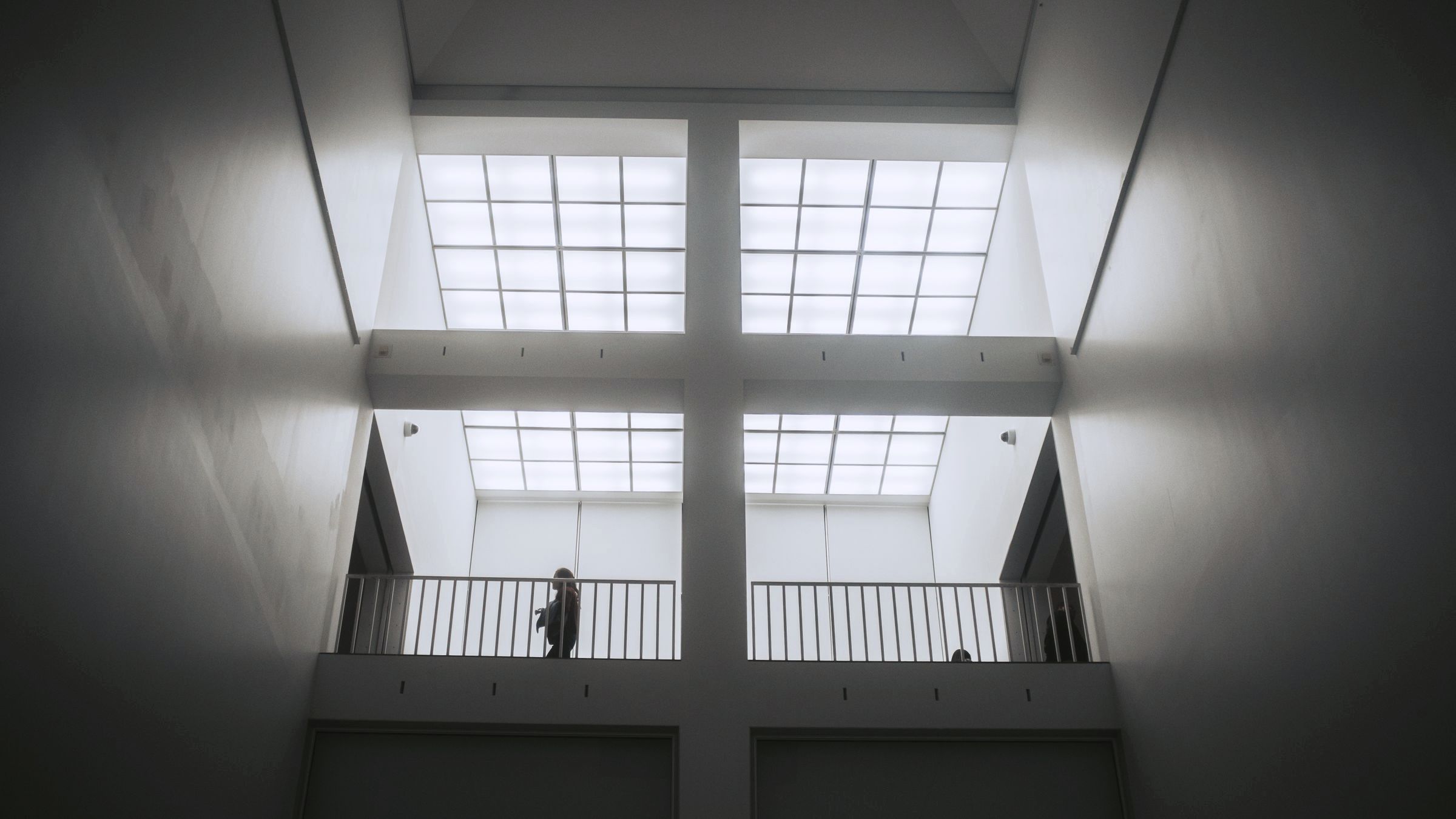 A white exhibition hall under a grid of light, a figure at the balustrade.