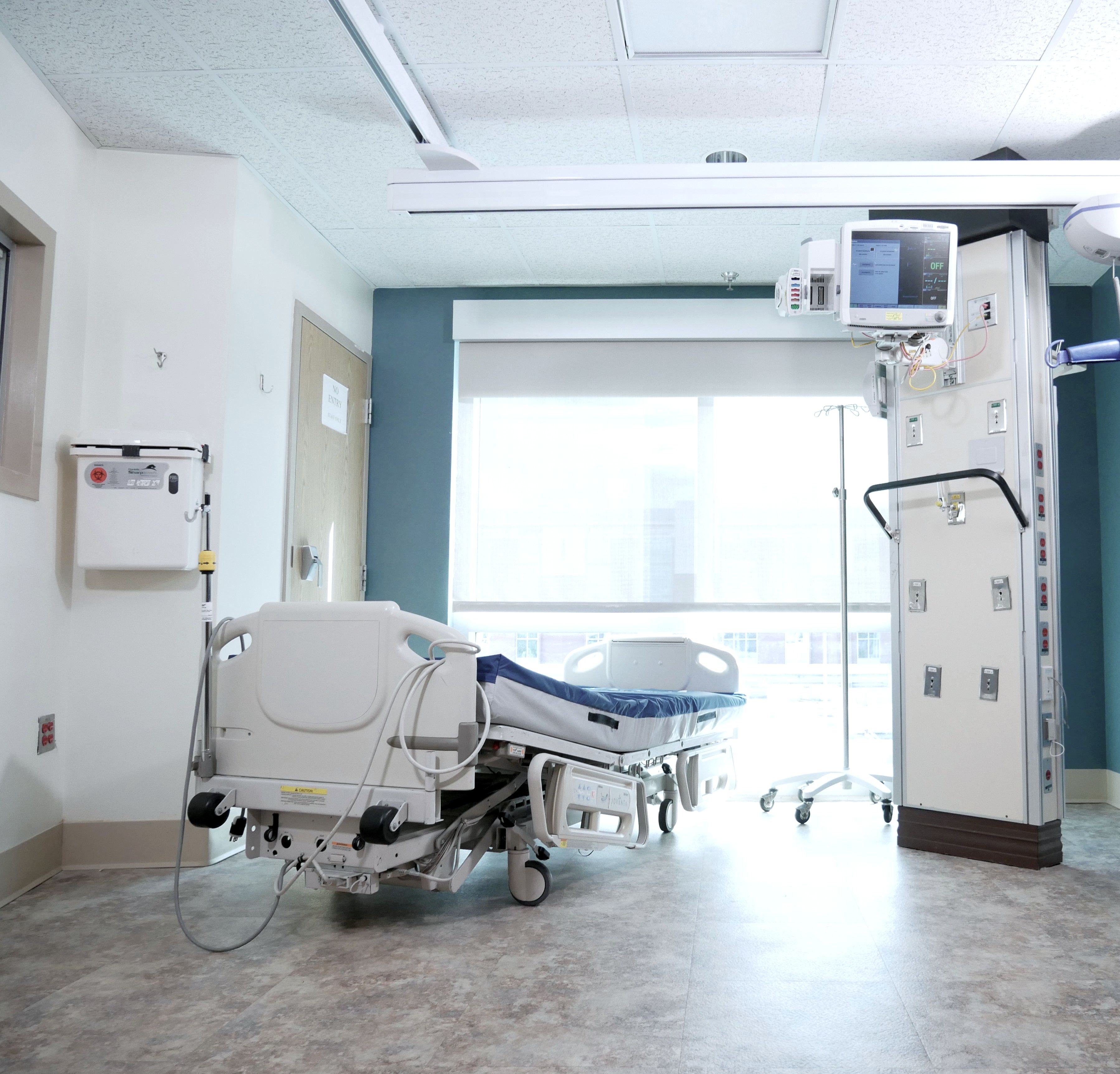An empty hospital bed in front of a large window, daylight falling across the room.