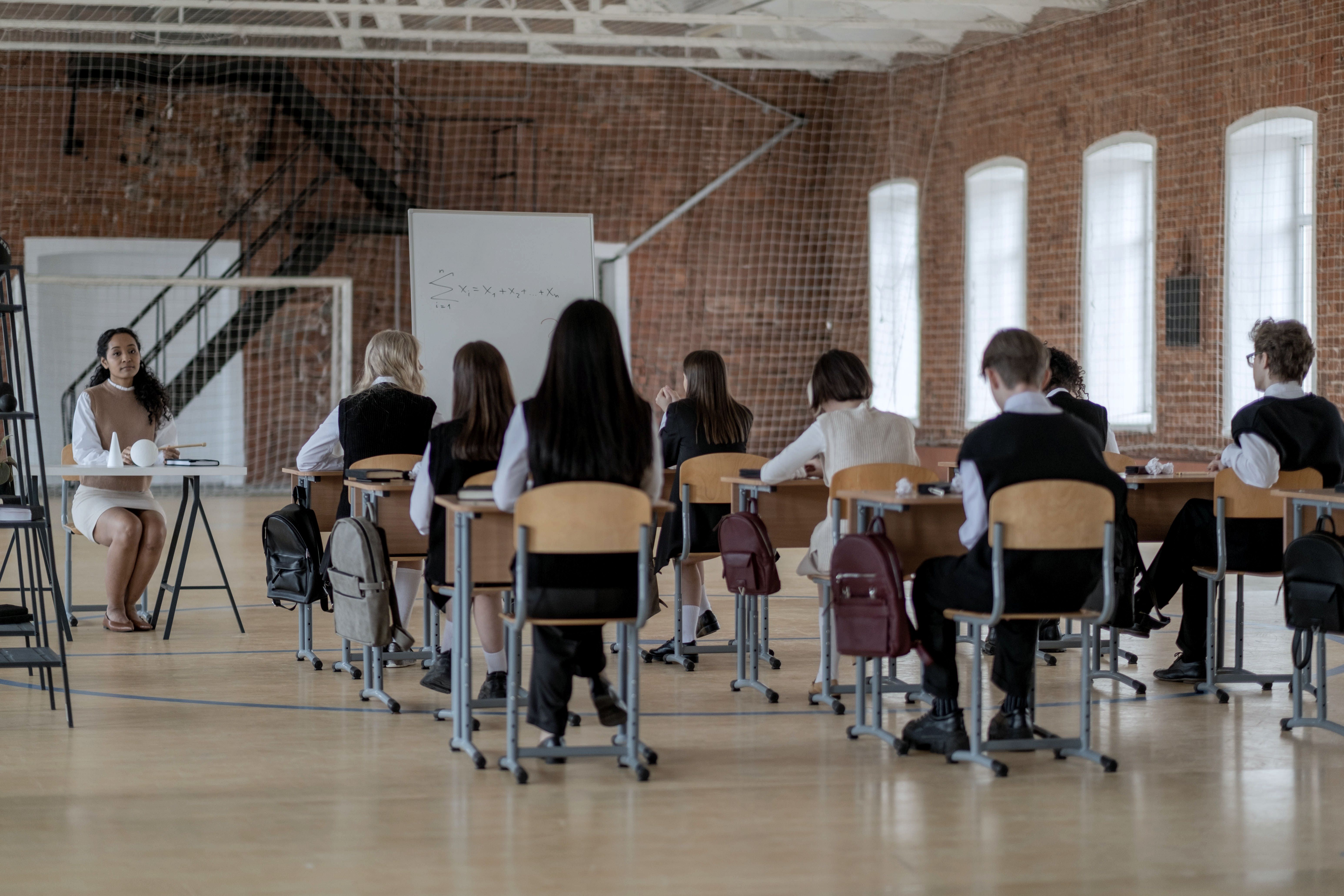 Pupils in rows seen from behind in a brick hall, daylight through tall arched windows.