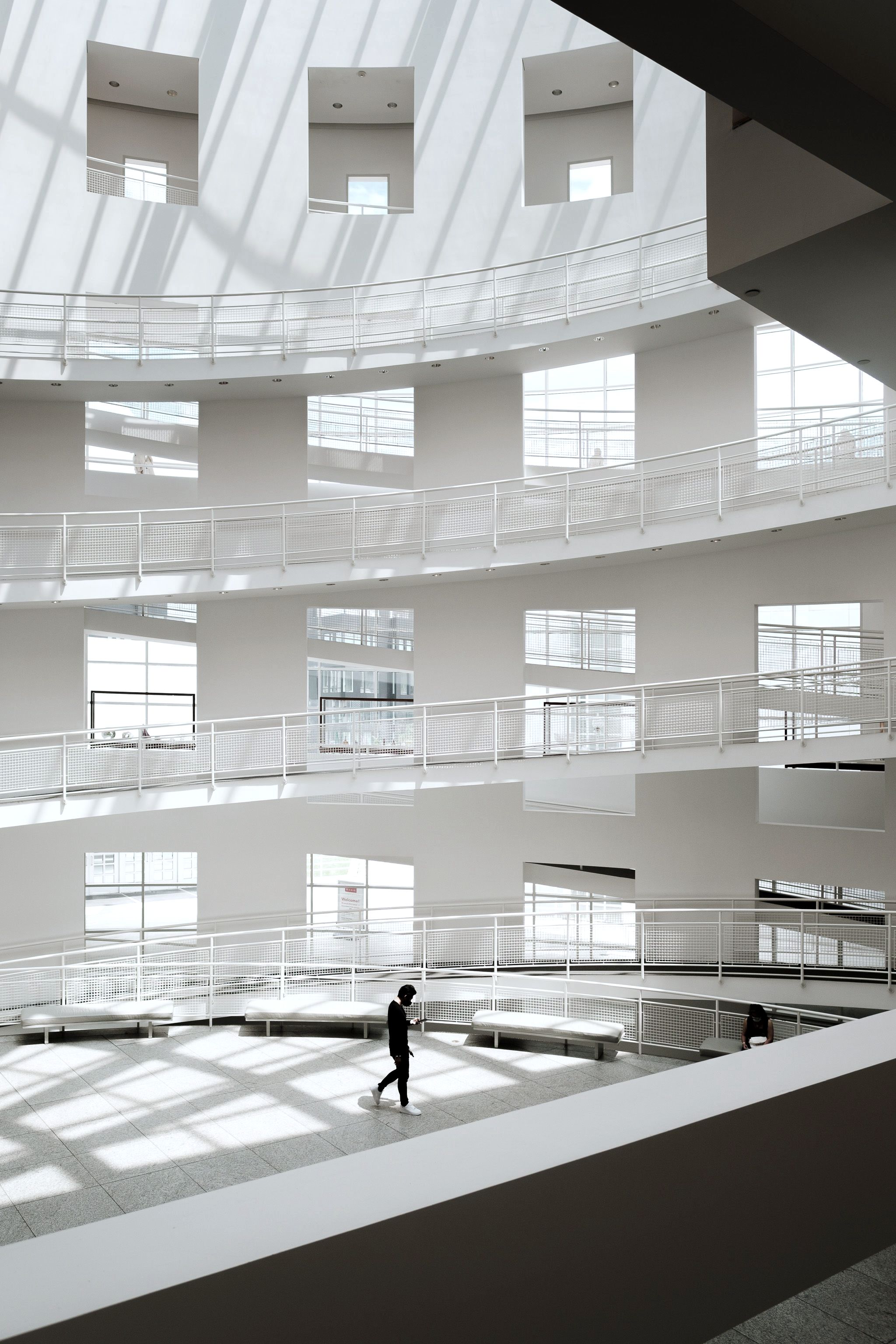 A white atrium of curved galleries, one person crossing the floor in low sunlight.