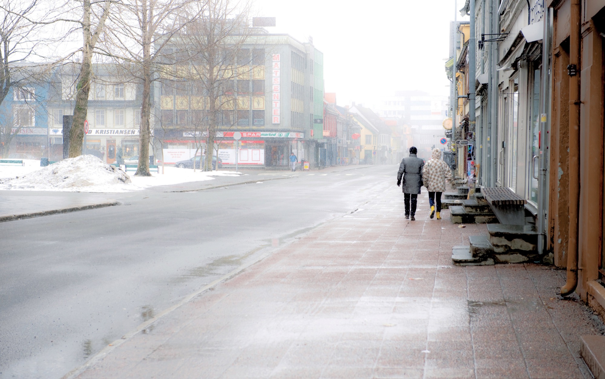 Two people walking along a quiet shopping street in light snow, in black and white