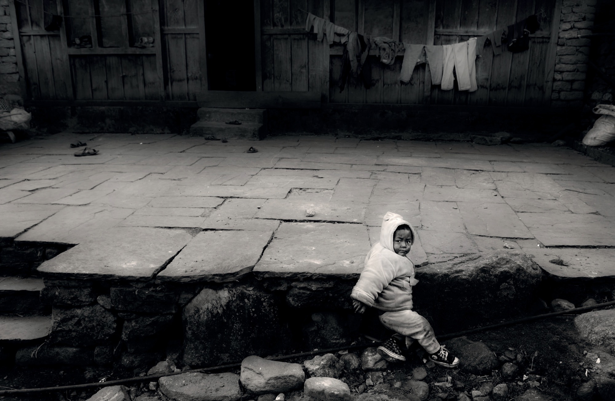 A child in a warm jacket sitting on a stone step in front of a wooden house, in black and white