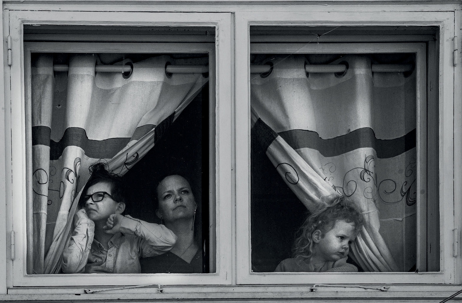 A woman and two children looking out of two windows between half-open curtains, in black and white