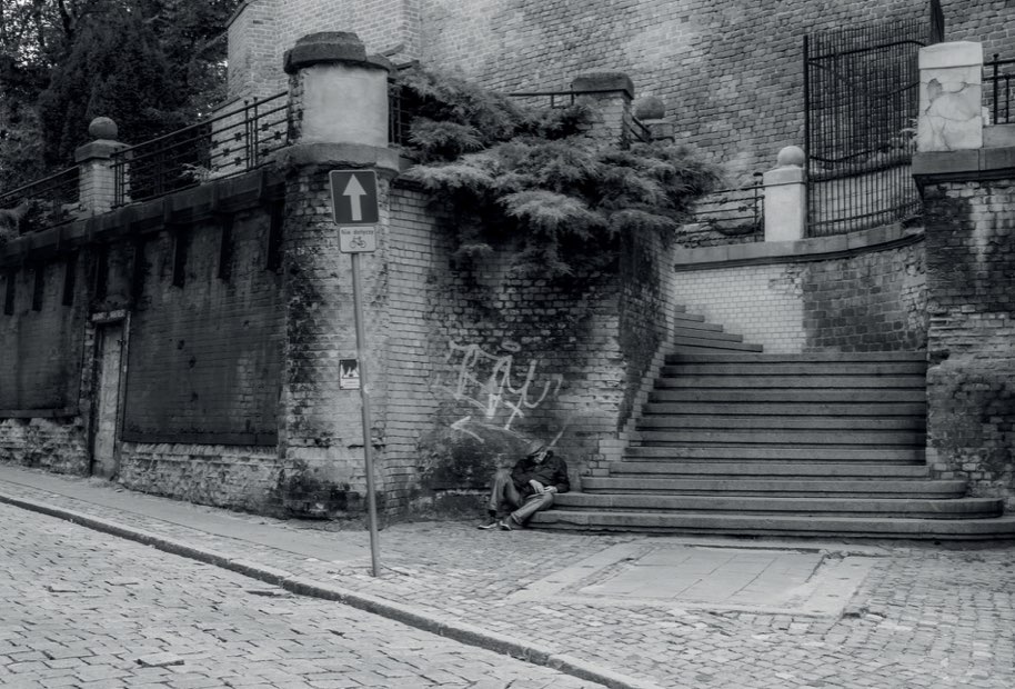 A man sitting alone at the foot of a stone staircase between brick walls, in black and white