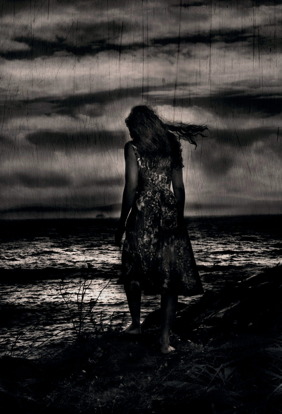 A woman in a dress standing at the edge of a dark sea under storm clouds, in black and white