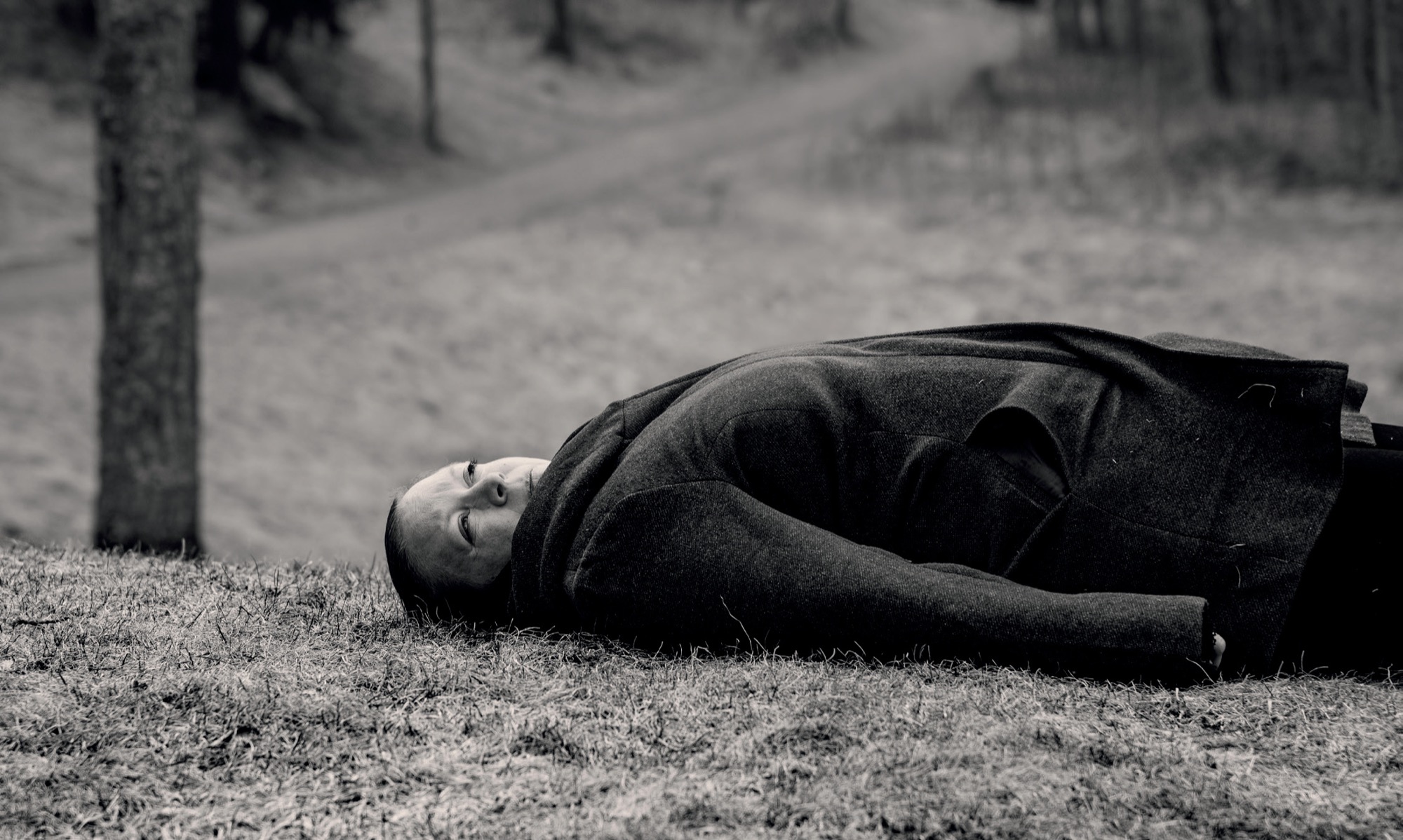 A woman in a dark coat lying on the ground in a forest clearing, in black and white