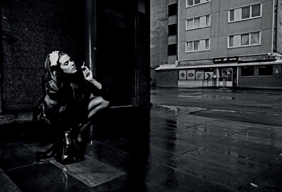 A woman crouching against a wall on a wet street corner at night, in black and white