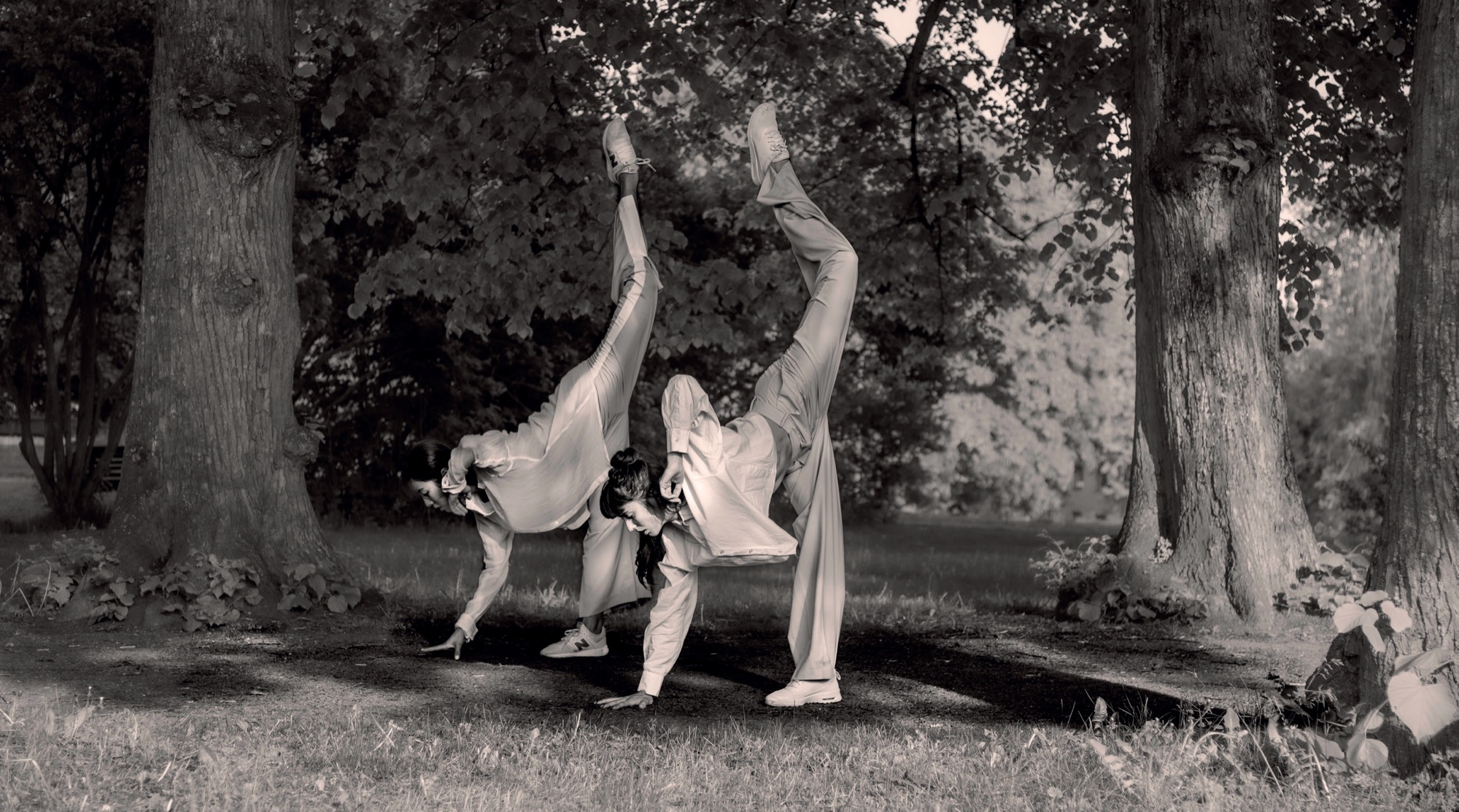 Two people doing handstands on a path between trees in a park, in black and white