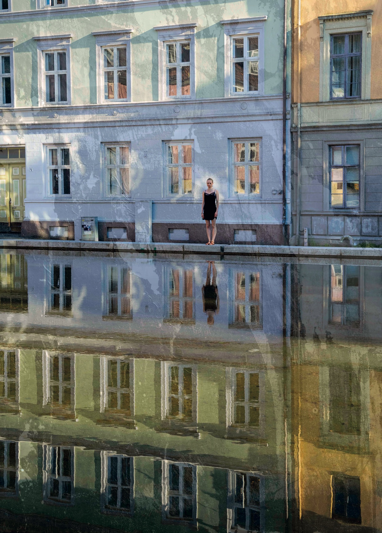 A young woman in a black dress standing at the edge of a still pool that reflects the house fronts behind her